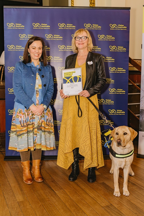 "Two women, one holding a certificate, and a seeing eye dog stand in a hall"